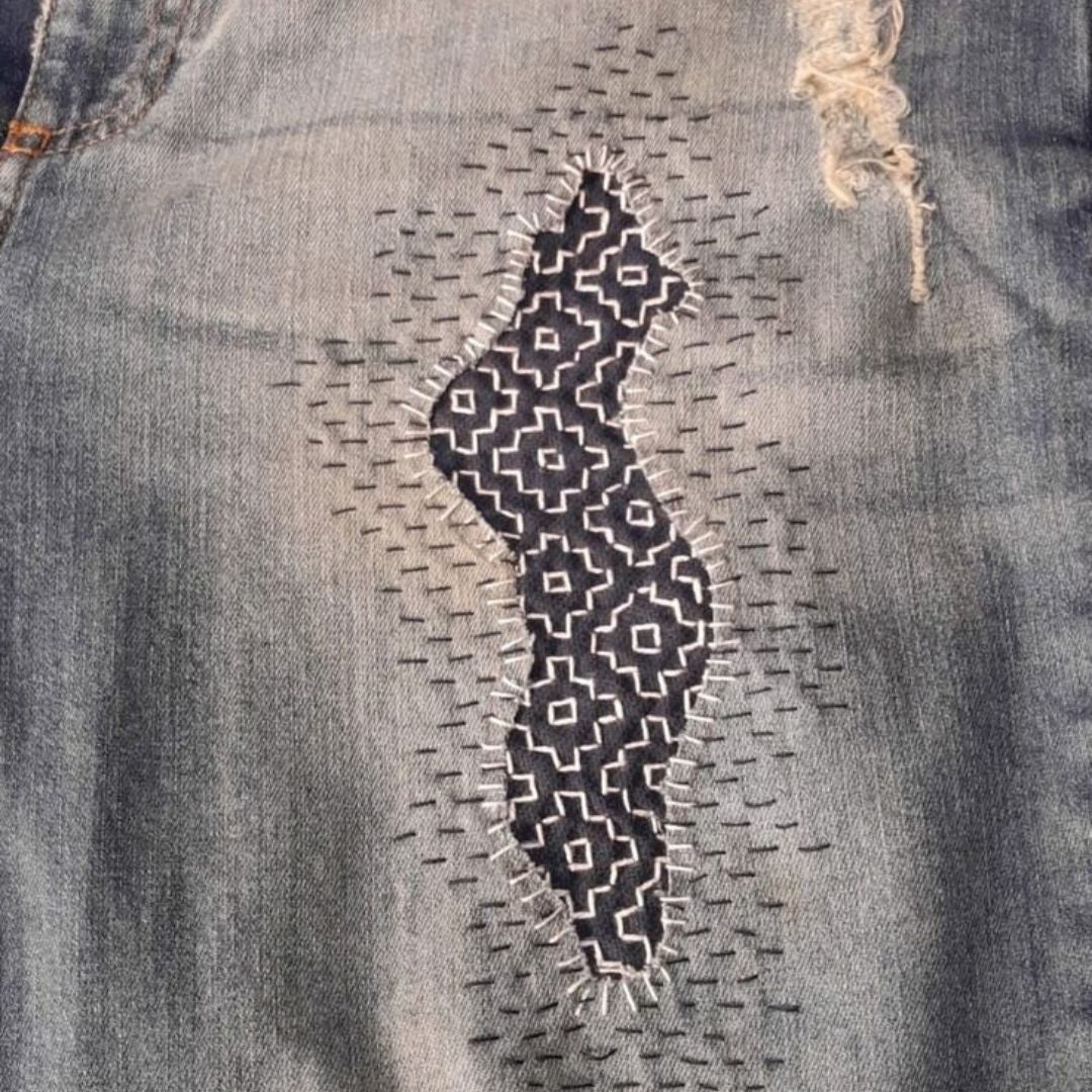 Close-up of worn denim repaired with visible hand stitching and patterned fabric, illustrating the craftsmanship of textile mending.